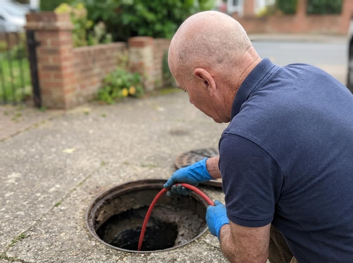 Philip rodding a blocked drain through an open manhole outside a house